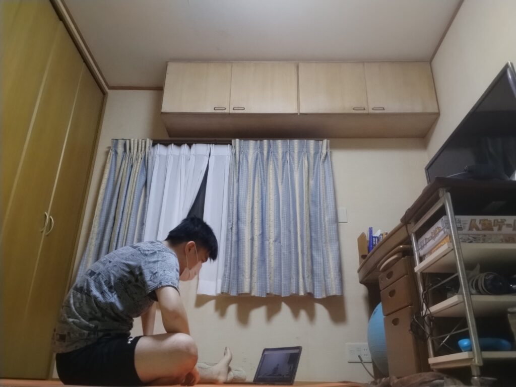 ノートパソコンの画面を見ながらヨガのオンラインレッスンを受講する男性。座位の姿勢でレッスン開始の準備をしている。