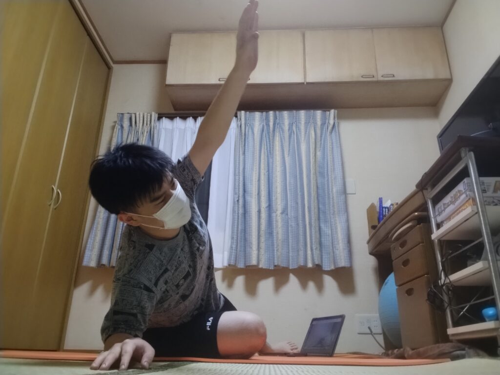 自宅でマスクを着けながらヨガのサイドストレッチポーズを行う男性。床に座り片腕を上げて体側を伸ばしている。