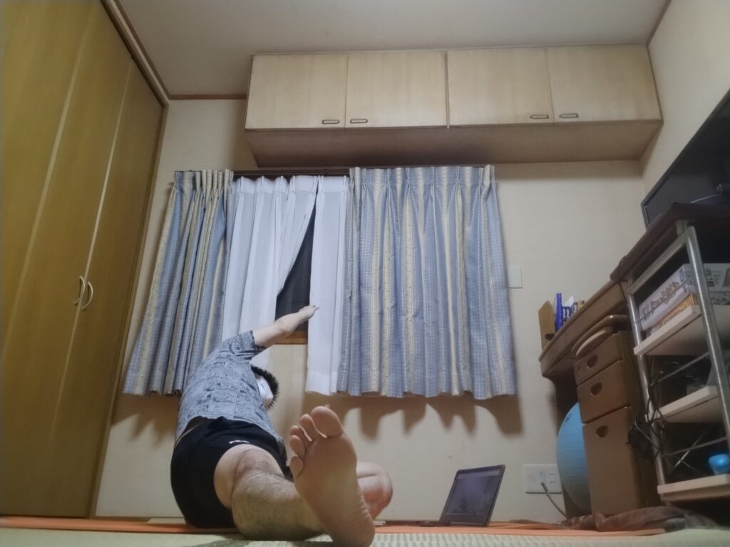 片脚を伸ばして座り、体側を大きく伸ばすストレッチをしている男性。オンラインヨガ中。