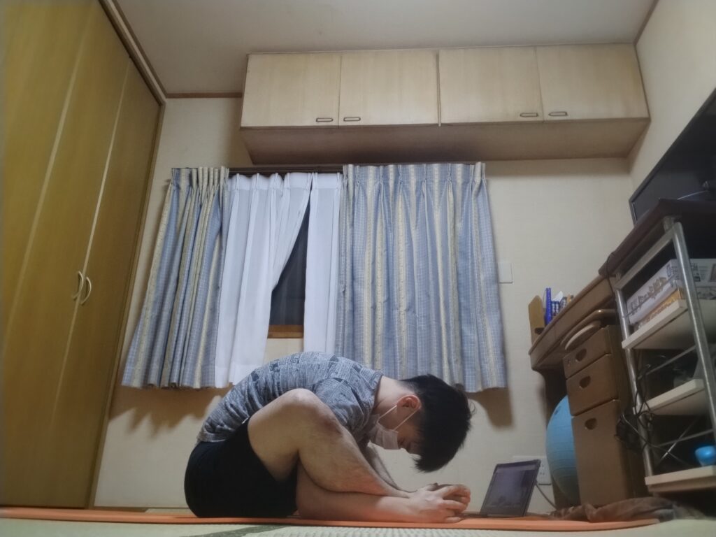 座位で前屈しながら足のつま先を持ち、太ももの裏や腰をストレッチしている男性。オンラインヨガのレッスン中の様子。