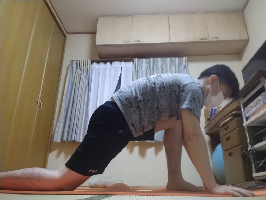 片膝立ちで股関節をストレッチするヨガポーズ。両手を床につけて体を支え、前脚は90度に曲げている。