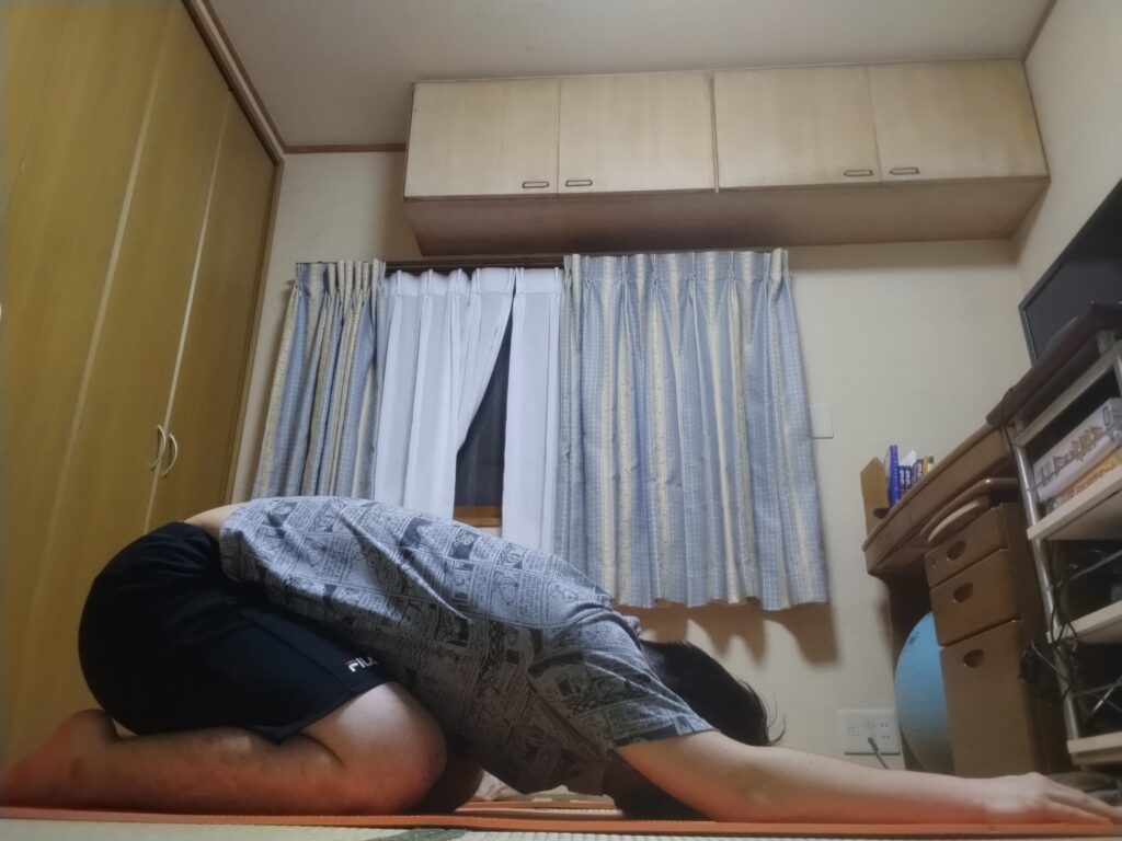 チャイルドポーズで床に伏せ、腕を前に伸ばしてリラックスしている男性。背中を丸めて深い呼吸をしている様子。