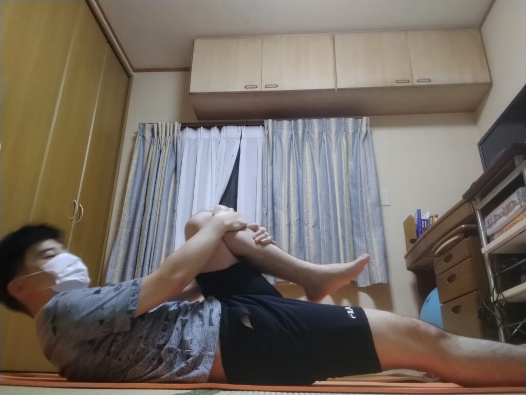 仰向けの姿勢から片膝を抱え、上半身を起こして体を丸めるストレッチをしている男性。体幹を使いながら腰と背中を伸ばしている様子。