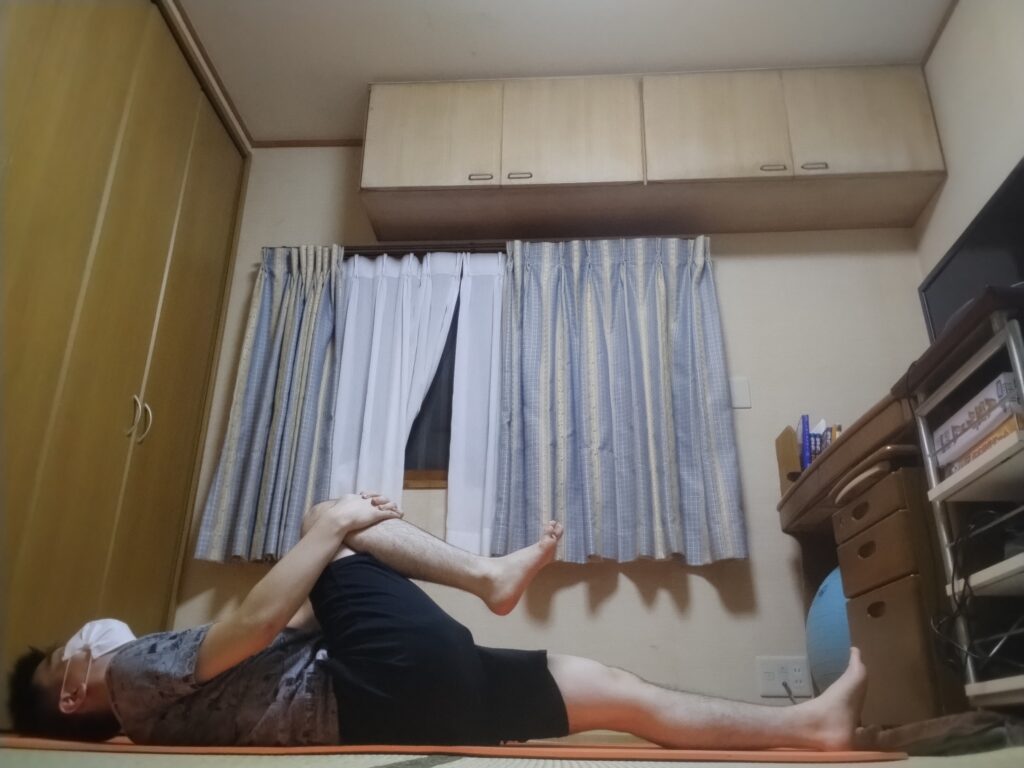 仰向けになり、片膝を胸に引き寄せてストレッチしている男性。