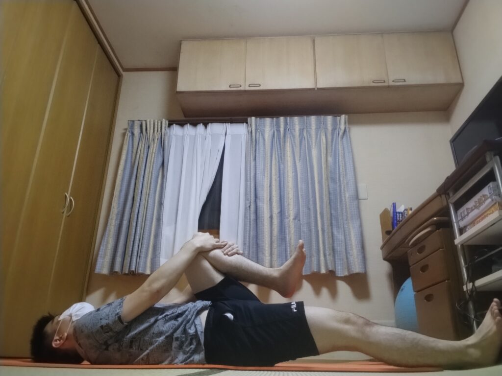 ヨガマットの上で仰向けになり、片膝を胸に引き寄せている男性。